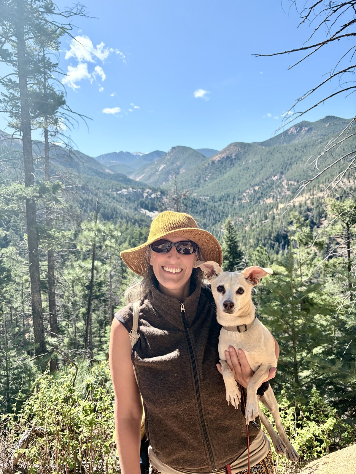 Connie in a straw sun hat holding Sami on a mountain trail with forested peaks behind them